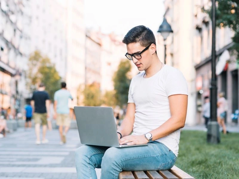 Młody mężczyzna szuka chwilówki od 18 lat siedząc na ławce z laptopem na kolanach.