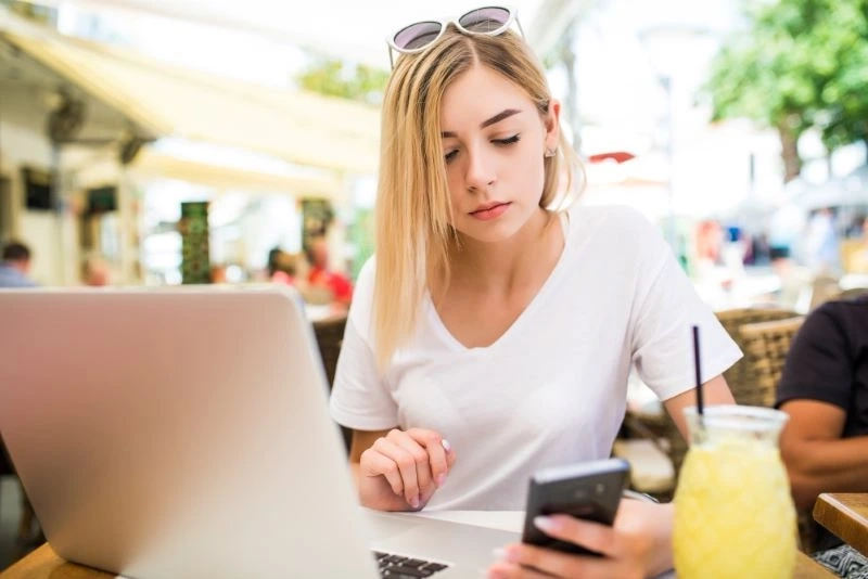 Młoda dziewczyna ze smartfonem szuka internetowej pożyczki od 18 lat siedząc z laptopem w kawiarni.